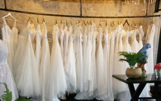 Reihe von Hochzeitskleidern an Metallständern in einem Geschäft, detailliert gezeigt mit einem Fokus auf die Spitzen- und Stoffverzierungen, vor einer Wand mit rustikalen und modernen Elementen