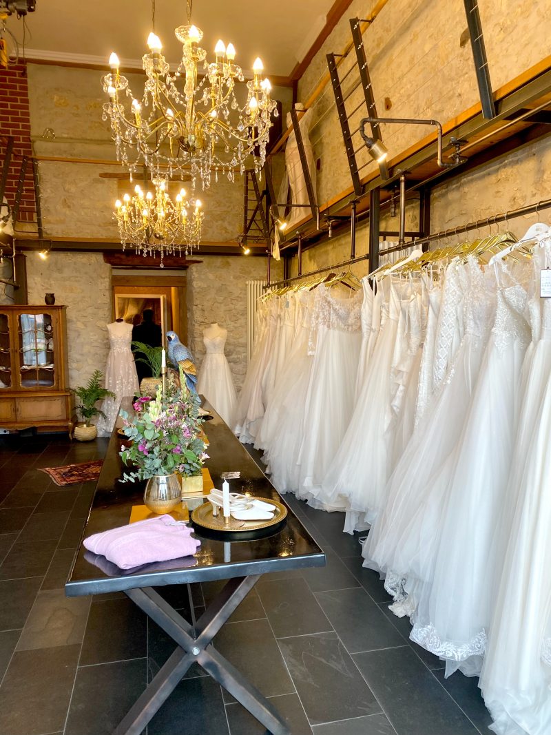 Ansicht eines Brautmodengeschäfts mit mehreren Kronleuchtern, einem langen, glänzenden schwarzen Tisch mit Hochzeitsaccessoires und Hochzeitskleidern an den Seiten, eingebettet in eine Umgebung mit rustikalen Steinwänden.