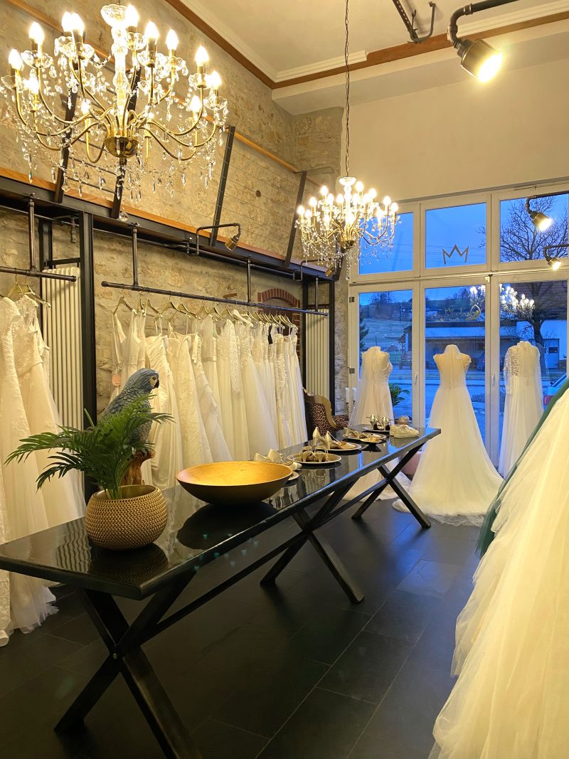 Ansicht eines Brautmodengeschäfts mit mehreren Kronleuchtern, einem langen, glänzenden schwarzen Tisch mit Hochzeitsaccessoires und Hochzeitskleidern an den Seiten, eingebettet in eine Umgebung mit rustikalen Steinwänden.